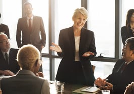A woman presenting in front of a table full of people, informally