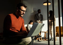 Mid adult man, sitting in a open space office, working on laptop computer, having online meeting while his colleagues have brainstorming in the background