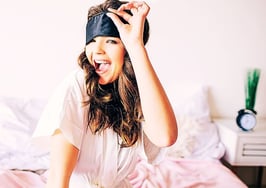 A woman in PJs in her bed pulling off a sleep mask to signify how top real estate agents start the day.