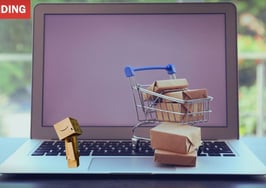 A small figure made of Amazon boxes looks at a computer behind a shopping cart full of packages, illustrating real estate data privacy.