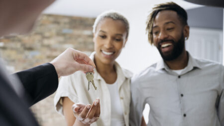 Over the shoulder view of real estate agent performing symbolic gesture of handing house keys to smiling homeowners in celebration of their purchase.