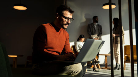 Mid adult man, sitting in a open space office, working on laptop computer, having online meeting while his colleagues have brainstorming in the background