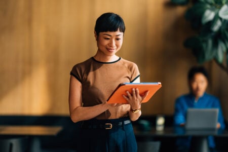 A confident woman using a digital tablet in a contemporary office environment. The setting conveys professionalism, technology, and productivity with a focus on modern work practices.